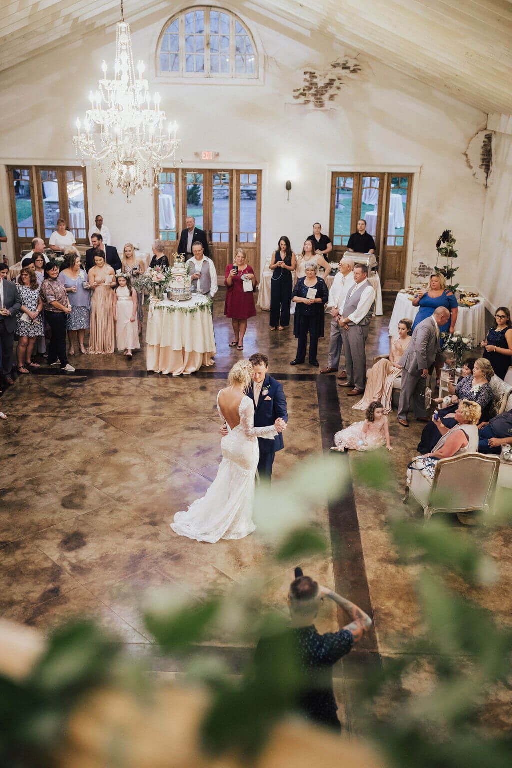 Bride and groom dance at reception; guests watch. Rustic venue with chandelier and arched window.