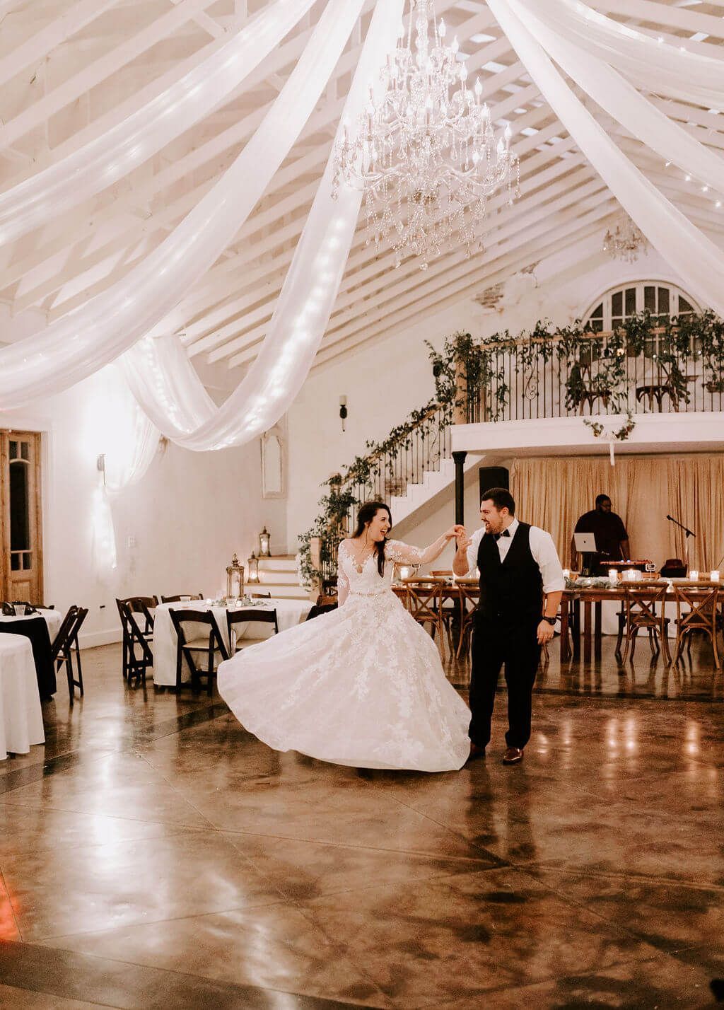 Couple dances in a decorated wedding venue with draping fabric and a chandelier.