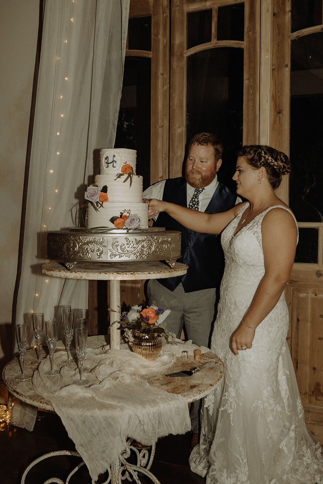 Bride and groom cutting wedding cake. Cake is white with floral accents on a tiered stand in front of a wooden door.