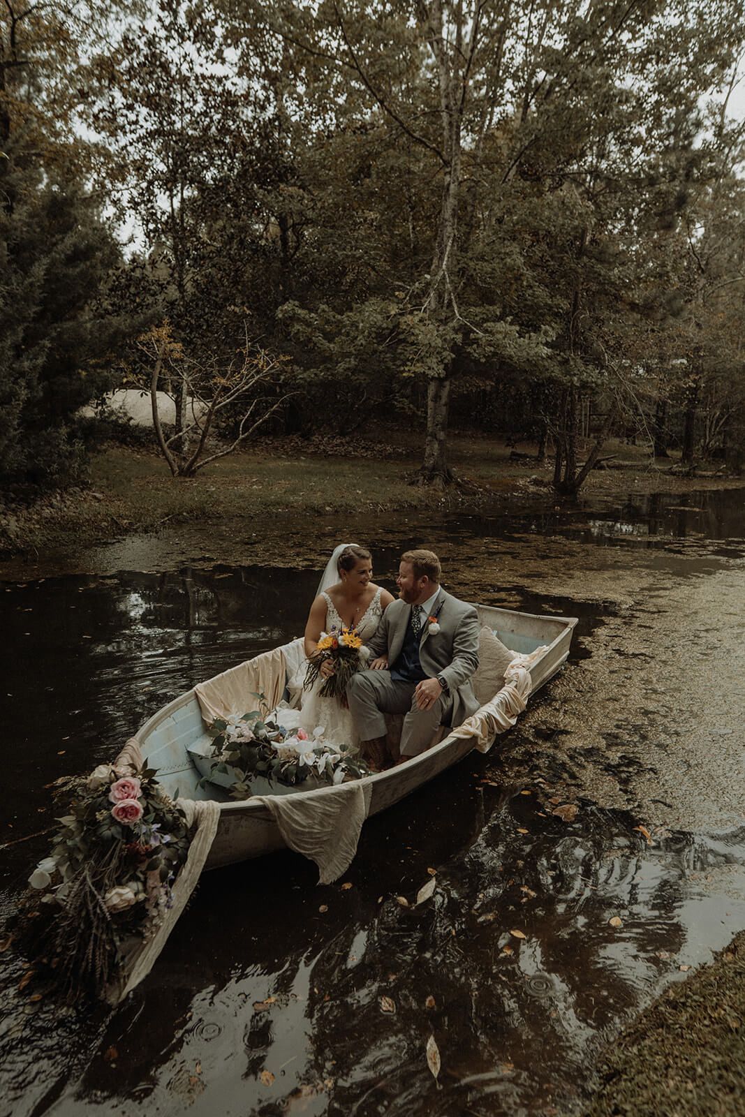 Couple in a decorated boat on a pond; bride in white dress, groom in suit, surrounded by trees.