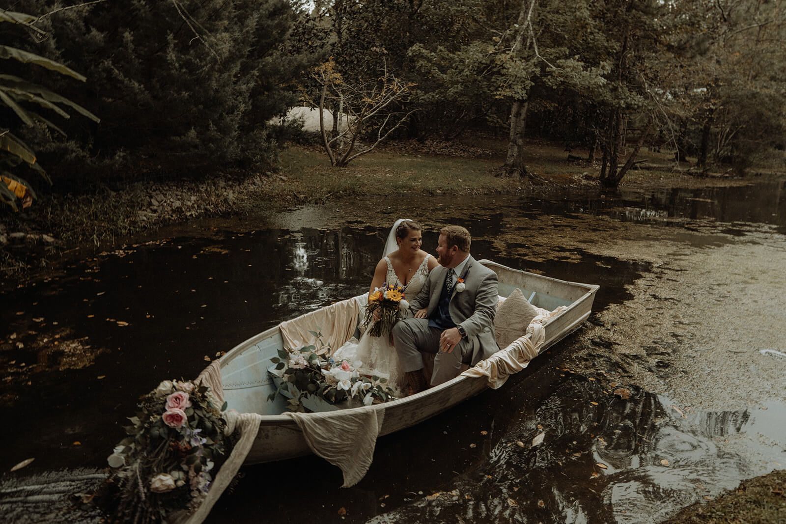 Newlyweds in a boat on a pond, surrounded by greenery. Bride in white dress, groom in gray suit.