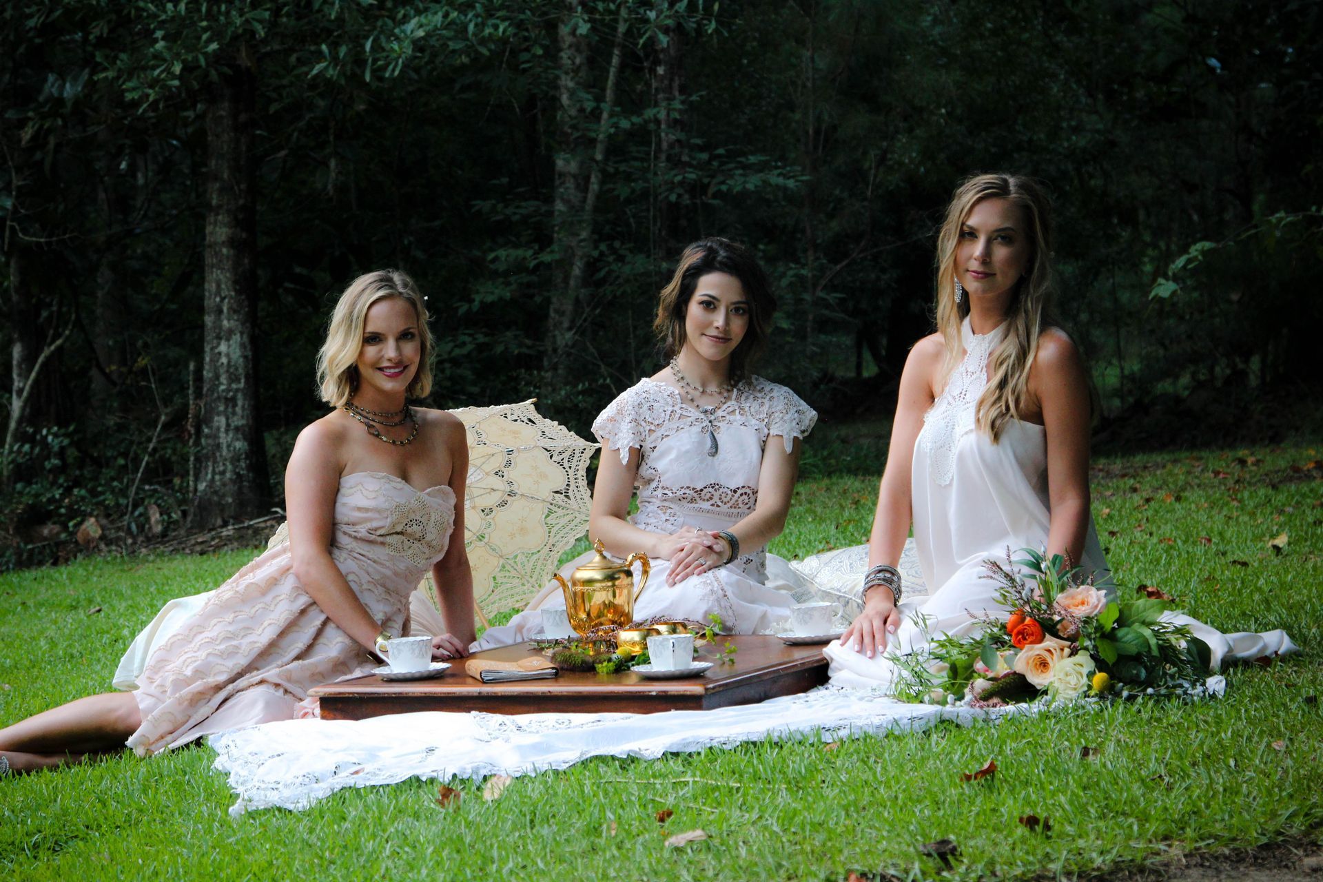 Three women having a picnic in a wooded area, sitting on a white blanket with food.