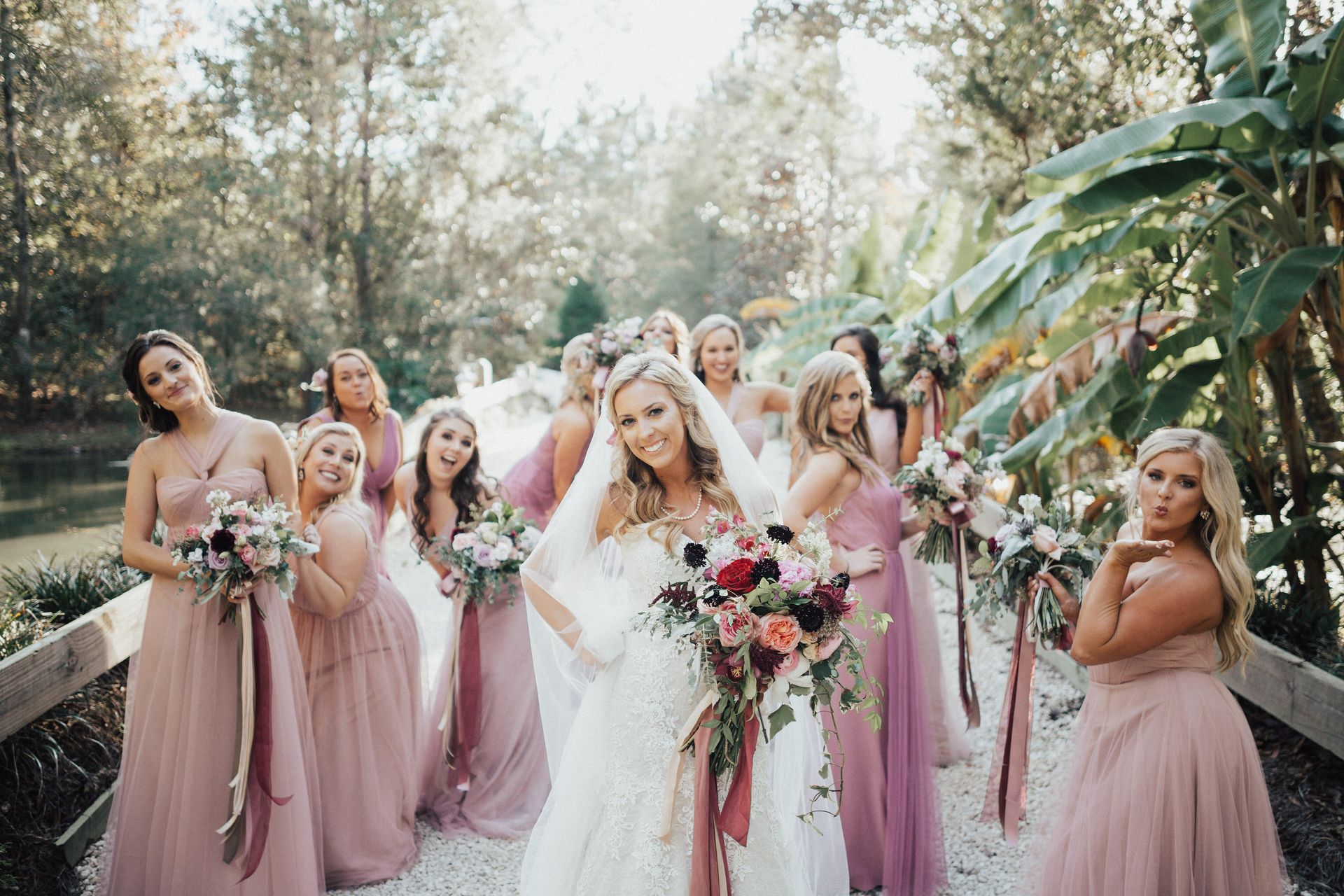 Bride in white gown poses with bridesmaids in pink dresses, outdoor setting.