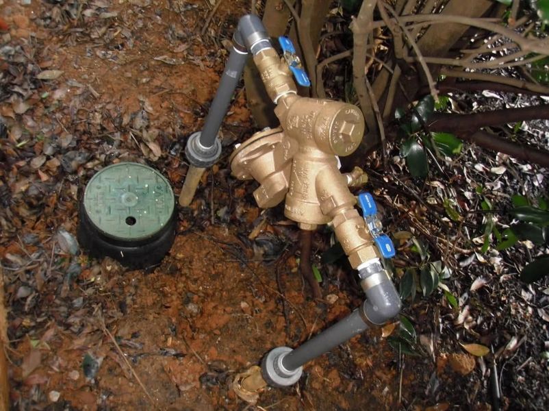 Backflow preventer and irrigation pipes beside a green water meter box in muddy ground