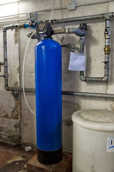 Blue water softener tank with plumbing pipes in a utility room next to a white container