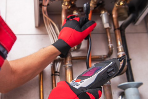 Gloved technician using a digital multimeter to inspect plumbing pipes under a sink