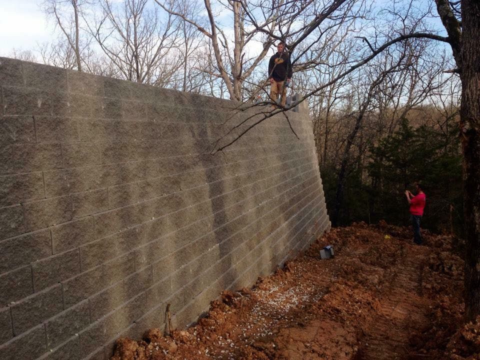 A tall, gray cinder block retaining wall with two people observing, one standing on it, in a wooded area.