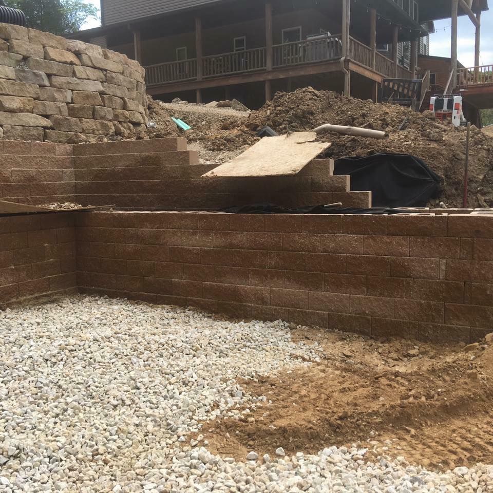 Stone retaining walls being built near a house; gravel base and dirt pile visible.