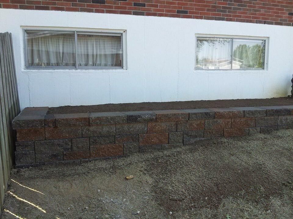 Brick retaining wall in front of white wall with windows; gravel ground.