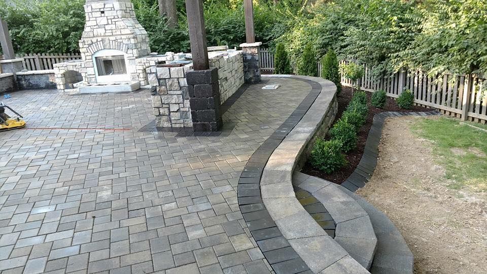 Brick patio with stone fireplace, curved walkway, and small shrubs in an outdoor setting.
