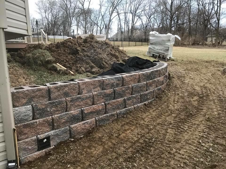 A retaining wall made of brown and gray blocks, next to a pile of dirt, on a grassy property.