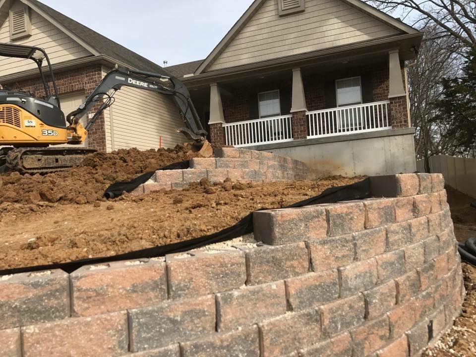 Excavator near house constructing a retaining wall of brown and red bricks. Earth on the ground.