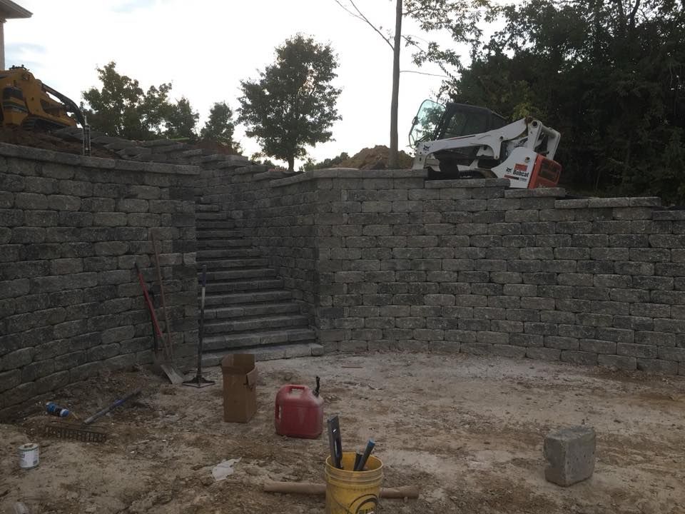 Stone retaining walls with stairs, construction site. Gray blocks, Bobcat, red gas can, and tools.