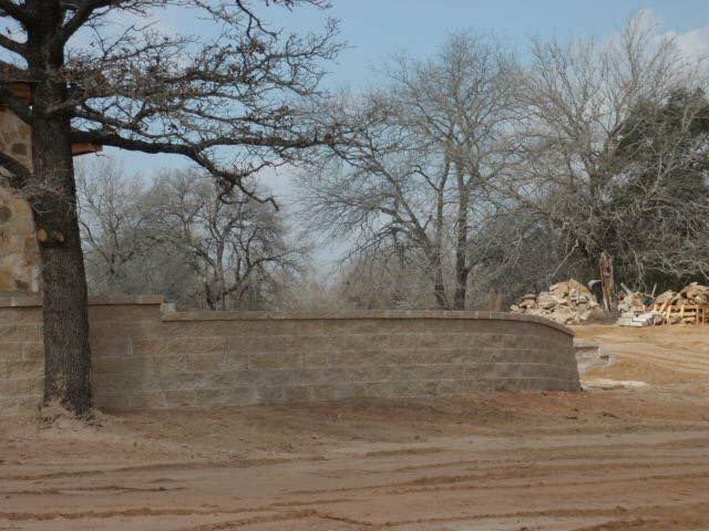 A tan stone wall curves in front of bare trees under a cloudy sky.