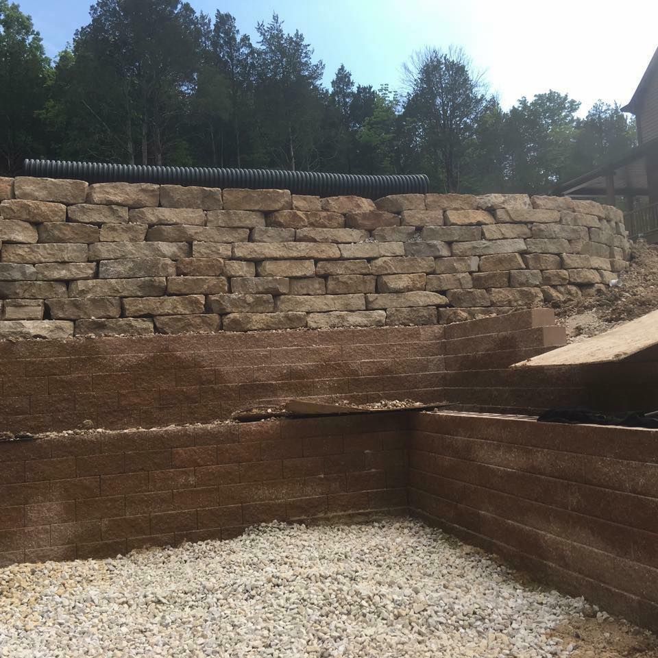 Stone retaining walls with a corrugated pipe above, gravel ground, and trees in the background.