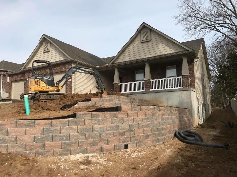 Excavator building brick retaining wall in front of a two-story house with a porch.