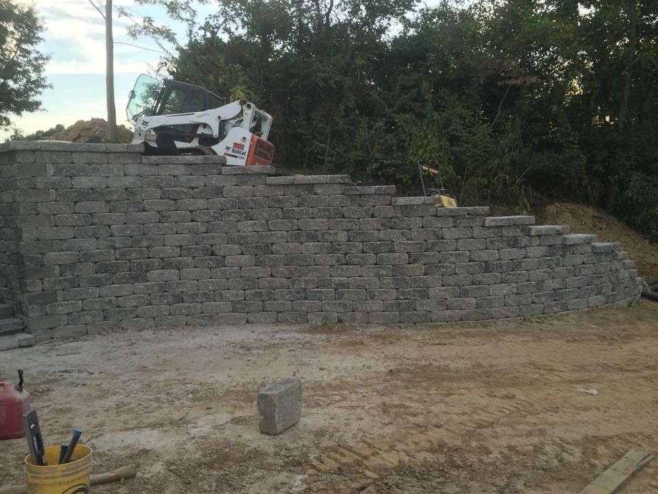 Gray retaining wall under construction with a Bobcat on top and tools in the foreground.