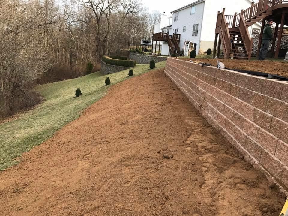 Retaining wall constructed on a slope next to a grassy area, with a house and trees in the background.