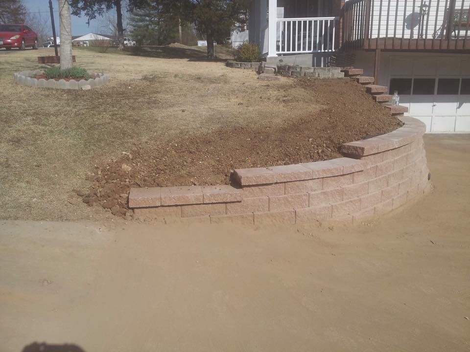 Brick retaining wall in front yard, with dirt and grass; driveway in the foreground.
