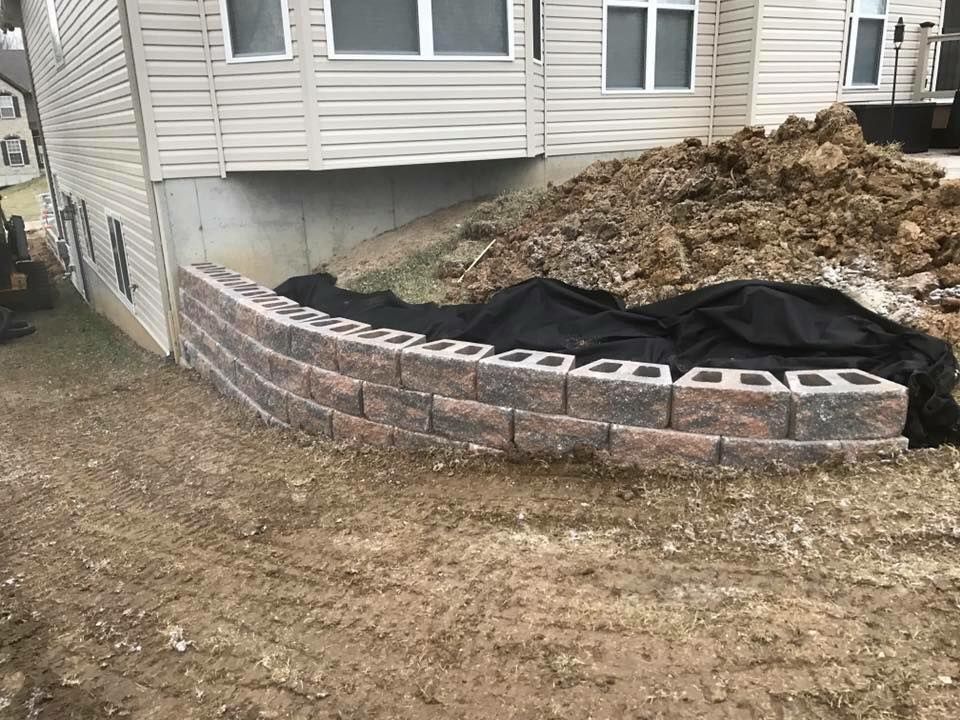 Curved retaining wall made of brown blocks, against a beige house. Soil and fabric visible behind the wall.