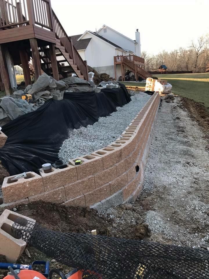 Stone retaining wall under construction near a deck and house. Gray gravel, black fabric.