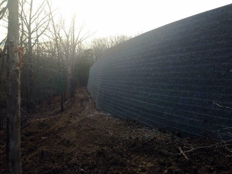 Dark, curved retaining wall made of wire mesh filled with dark rock, beside a wooded hillside.