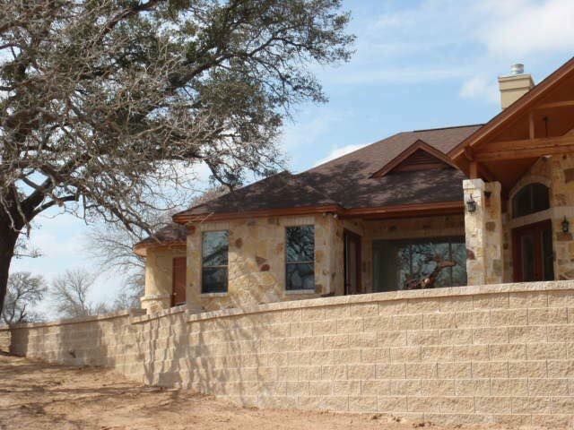 Beige stone house with brown roof and retaining wall, under a tree.
