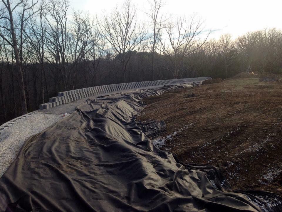 Black tarp covering a sloped embankment, with a retaining wall and trees in the background.