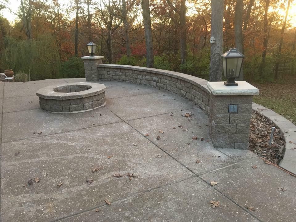 Stone fire pit and curved wall with lamps on a concrete patio. Forest background with fall foliage.