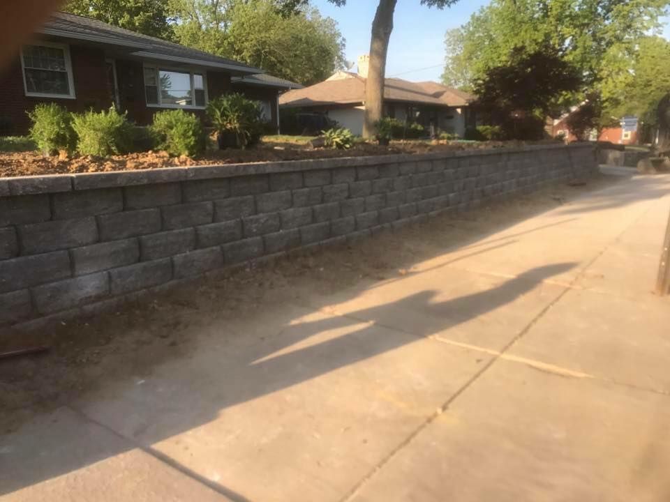 Gray retaining wall along a sidewalk, with houses in the background and the shadow of a tree.
