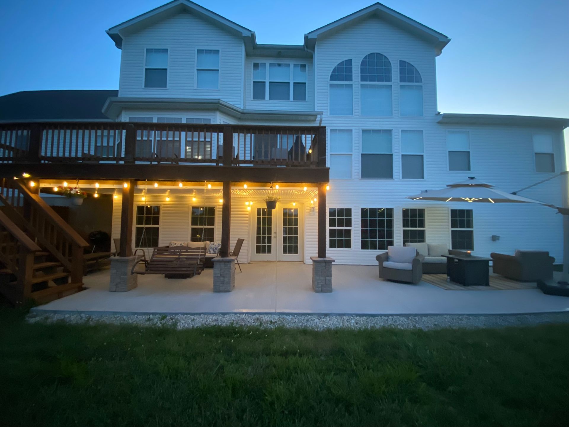 Backyard of a two-story white house with a deck, patio, outdoor furniture, and string lights at dusk.