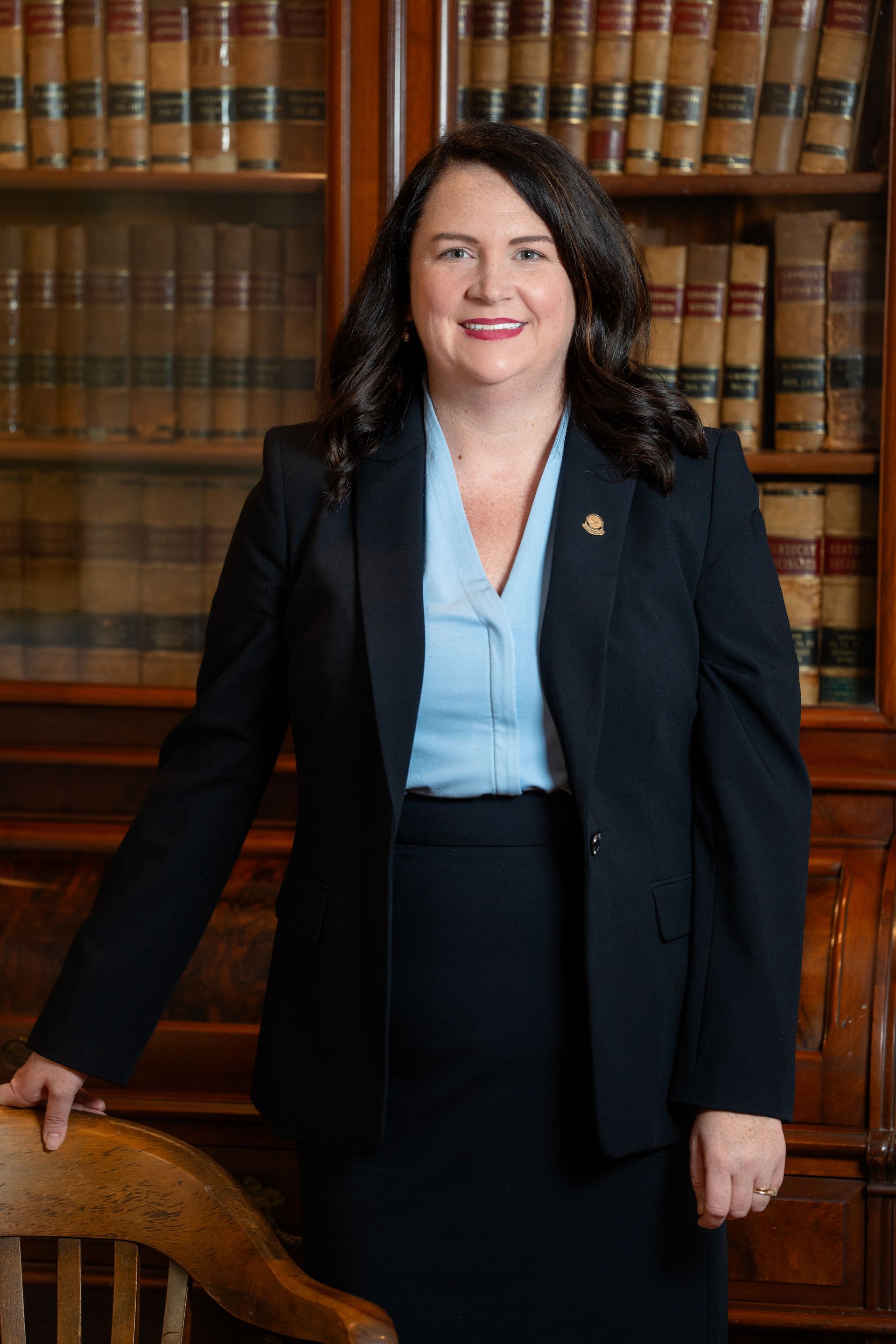Woman in a dark suit smiles, standing near a wooden desk and bookshelf.