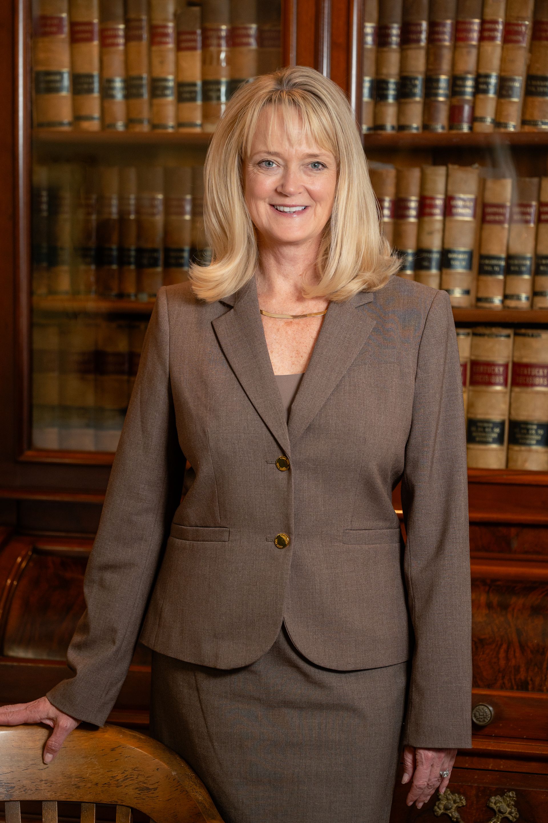 Woman in brown suit smiles, posed in front of a bookshelf filled with books.
