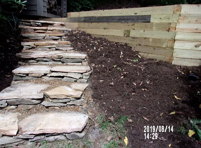 Stone steps alongside a fresh mound of dark mulch next to a light-colored wooden retaining wall in a backyard garden.
