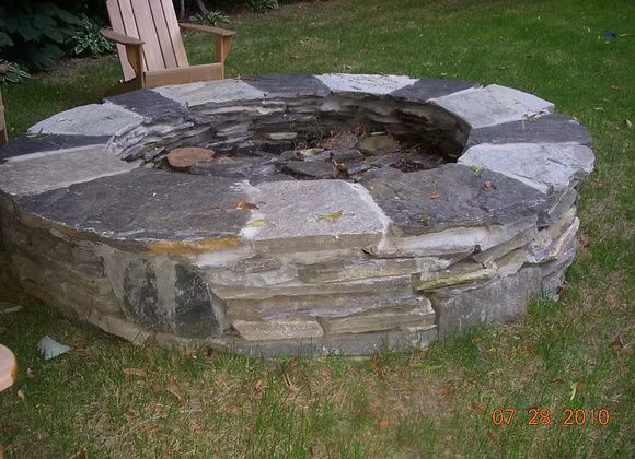 A circular stone fire pit sits in a grassy yard, topped with large, flat grey capstones.