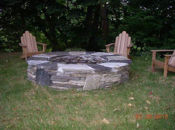 A stone fire pit surrounded by three wooden Adirondack chairs in a grassy backyard setting.