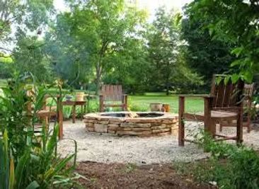 A circular stone fire pit sits in a gravel clearing surrounded by wooden chairs in a lush, green backyard.