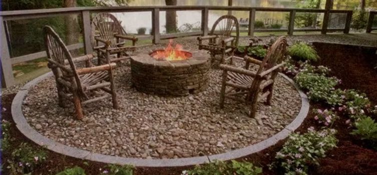 A stone fire pit with a lit fire, surrounded by four rustic wooden chairs on a circular gravel patio in a backyard.