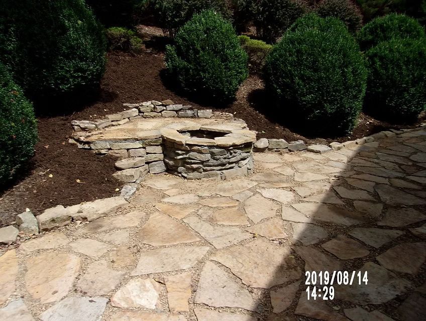 A stone patio with a built-in stone fire pit, surrounded by manicured shrubs and landscaping mulch.