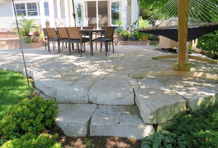 A stone patio with a dining table and chairs, featuring a stone step leading down to a green lawn with a hammock nearby.