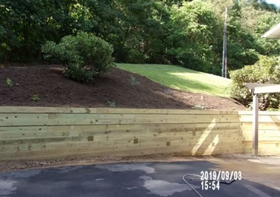A new wooden retaining wall borders a sloped garden bed covered in dark mulch with a grassy hill in the background.