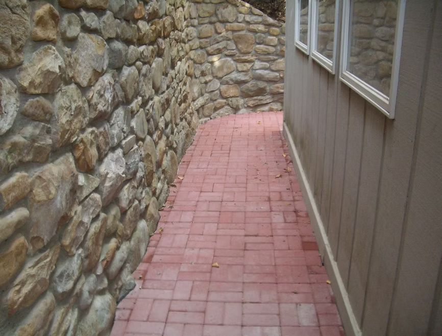 A narrow, paved red brick path runs between a rustic stone retaining wall and the vertical wood siding of a building.