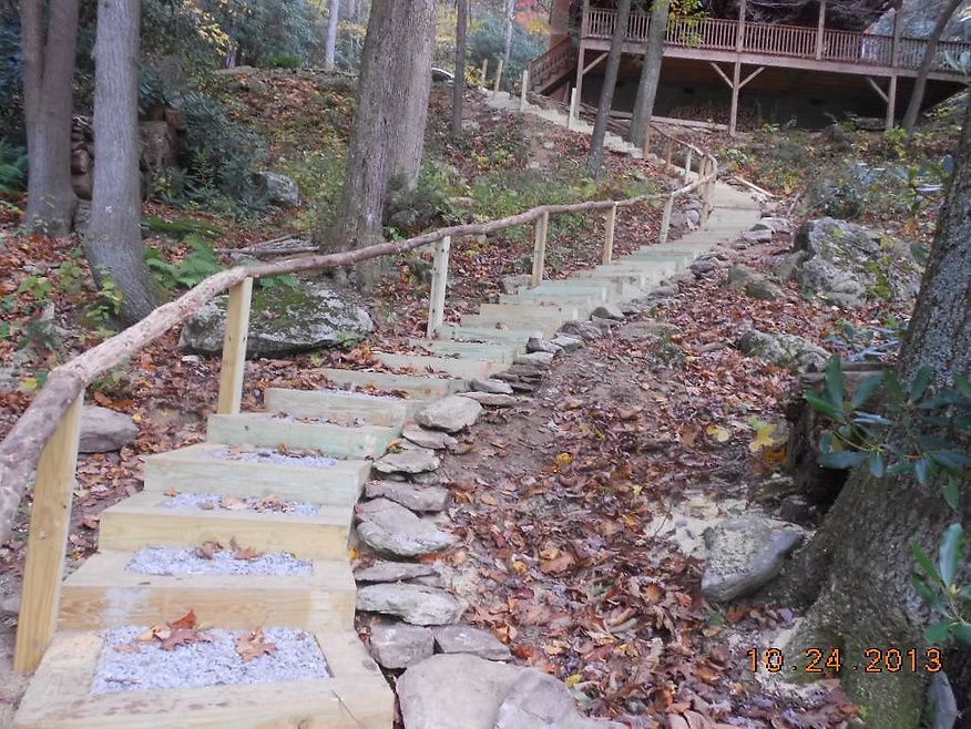 A wooden staircase with gravel steps winds up a leaf-covered, wooded hillside toward a house deck with a rustic railing.