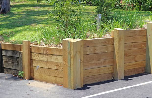 A wooden retaining wall constructed with thick sleepers and vertical support posts, separating a grassy area from a road.