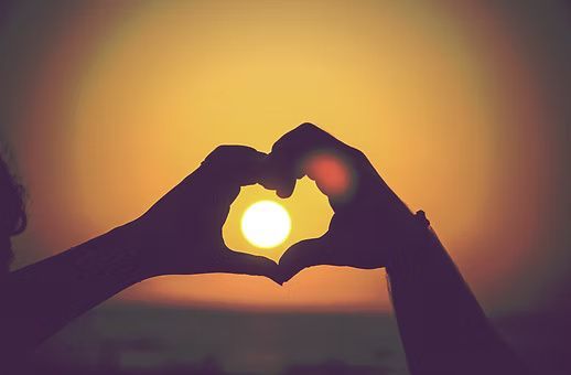 Silhouetted hands forming a heart shape around a bright, glowing sun against a golden sunset sky.