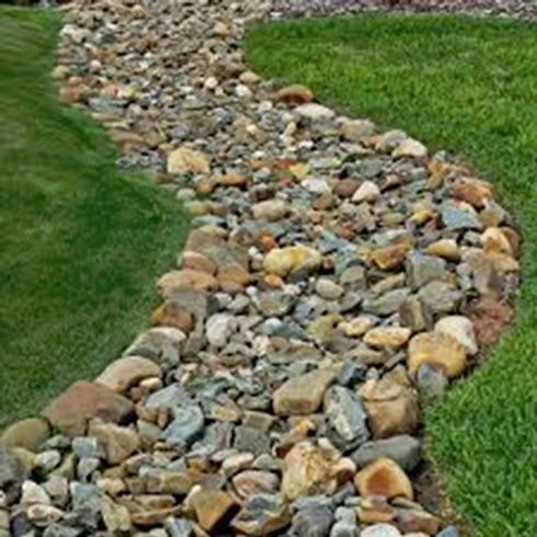 A winding dry creek bed made of various sized grey and brown stones, bordered by patches of green grass.