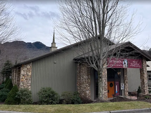 A brown, modern building with a parking lot in front sits before a large, green mountain range under a blue sky.
