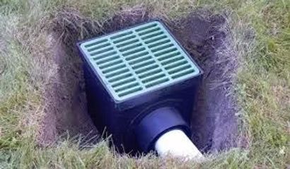 A square green drainage catch basin with a white pipe installed in a hole in the ground surrounded by grass.