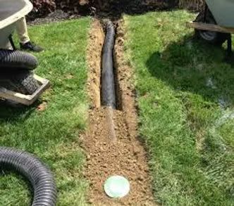 A black corrugated drainage pipe lies in a narrow trench dug through a grassy lawn, with a round green cover nearby.