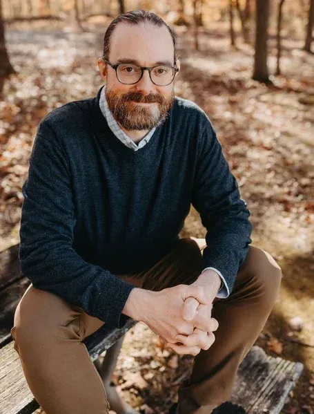 Man with glasses and beard, in a dark blue sweater, sitting outdoors on a bench, hands clasped.